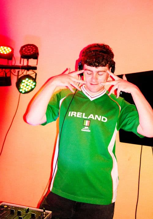 Person wearing green Ireland jersey and headphones, posing near DJ equipment with colorful stage lights.