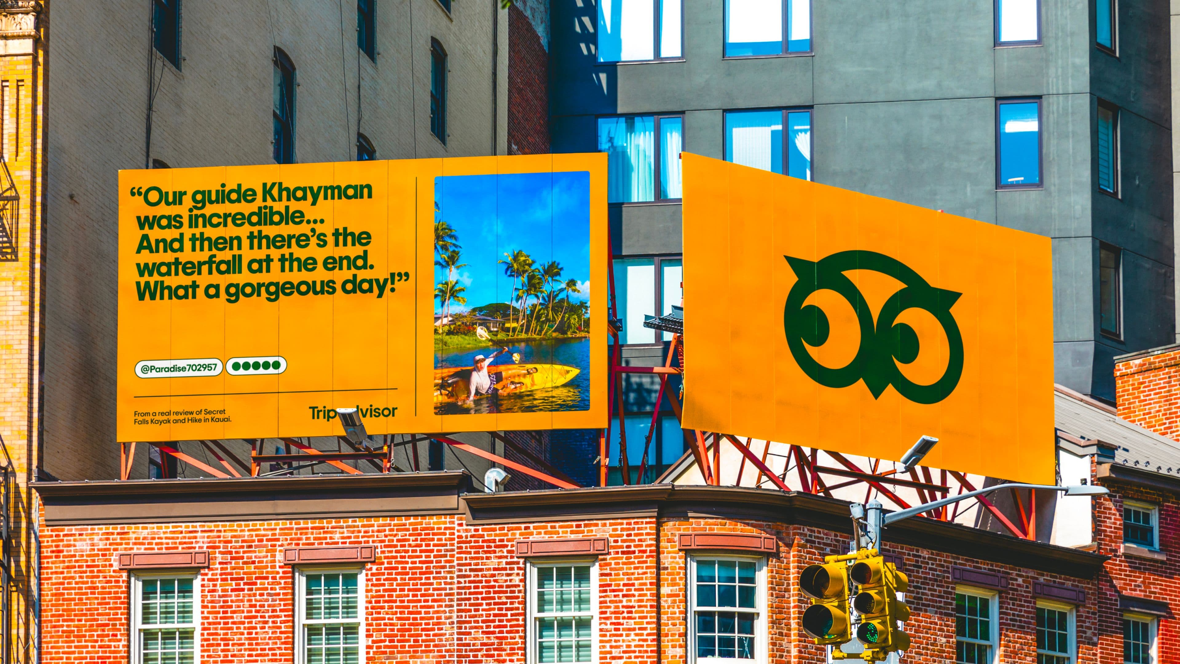 Two bright yellow TripAdvisor billboards above city buildings: one shows a review with a photo of two people by a tropical waterfall, and the other features the TripAdvisor owl logo.