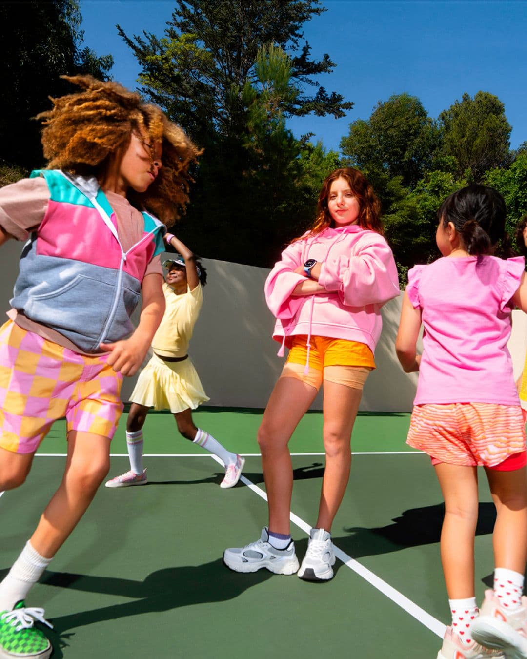 Girls playing and dancing on an outdoor court, colorful outfits, sunny day; center girl in pink wearing an Ace Fitbit.